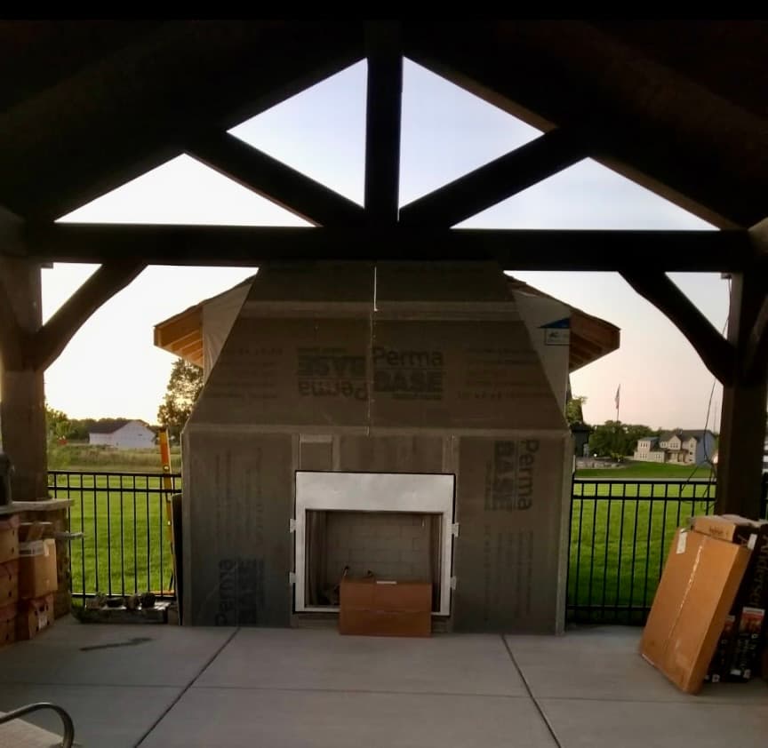 Masonry outdoor fireplace being built under a timber-framed pavilion overlooking a backyard